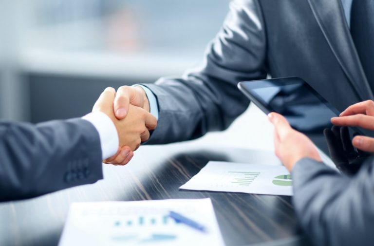 Two businesspeople shake hands while a third taps a tablet