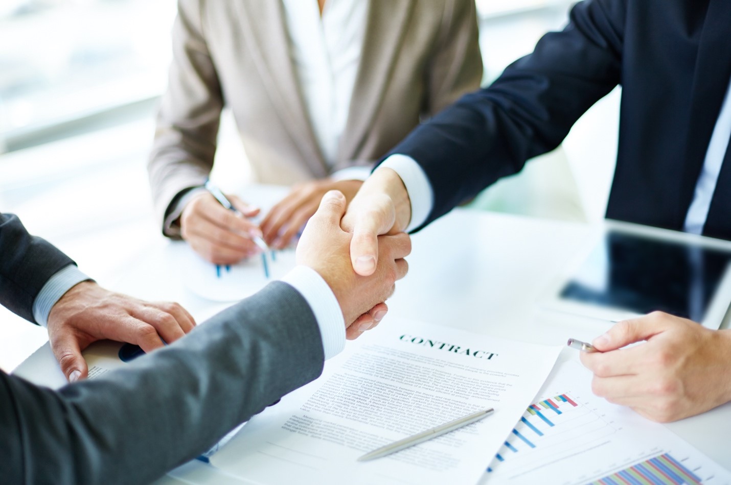 Two businesspeople shake hands over a contract while an attorney looks on
