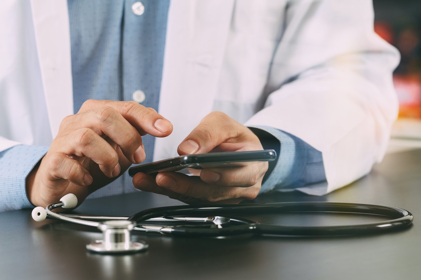 Closeup of doctor in white lab coat using a smartphone next to a desk with stethoscope.