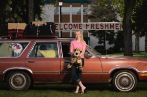 A young woman in cuffed jeans holding a stuffed animal beside a vintage station wagon. A large banner in the background reads WELCOME FRESHMEN